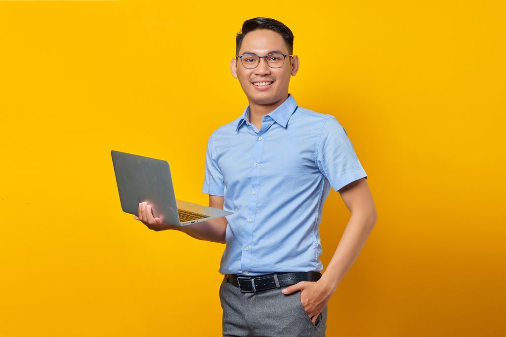 Modern Workplace A confident young adult male with eyeglasses, holding a laptop against a vibrant yellow background, exuding a professional vibe.